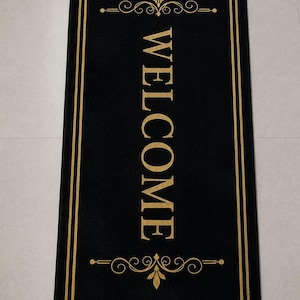 May include: A rectangular black floor mat featuring a gold-coloured border and the word WELCOME printed vertically in gold capital letters. The design includes gold decorative scrollwork accents above and below the text on the black surface.
