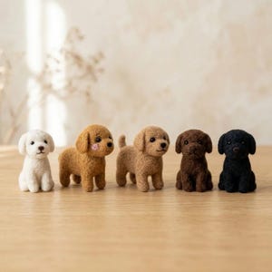 以下が含まれることがあります： 一列に並んだ5体の小さな羊毛フェルトの犬のフィギュア。白い犬、頬にピンクのアクセントがあるライトブラウンの犬、ミディアムブラウンの犬、ダークブラウンの犬、黒い犬のコレクションで、すべて黒いビーズの目と鼻が付いています。