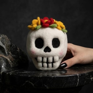 May include: A needle-felted white skull figurine featuring black eye sockets and a black nose cavity. The skull has a row of white teeth and soft pink blush on the cheeks. A crown of yellow and red felted flowers sits atop the skull head.