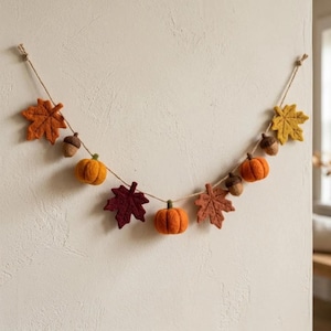 May include: A felt garland featuring orange, yellow, and dark red maple leaves, small orange pumpkins, and brown acorns strung along a thin twine cord. The decorative piece displays a variety of autumn-themed shapes in soft textures.