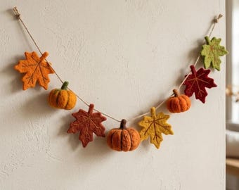 May include: A felt garland featuring orange, red, and yellow maple leaves interspersed with small orange pumpkins. The felt shapes are strung along a thin, natural-colored twine, creating a decorative hanging piece for seasonal display.