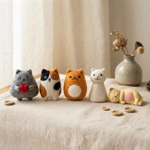 Peut inclure: Cinq figurines de chat en laine feutrée disposées en rangée. Le premier est gris tenant un cœur rouge, le deuxième est tricolore, le troisième est orange avec un ventre blanc, le quatrième est blanc et le cinquième est un chat jaune couché.