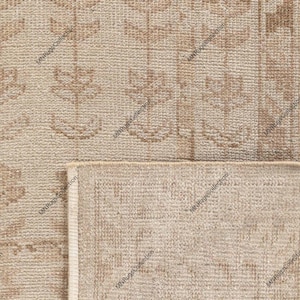 May include: A woven rug featuring a light beige background with a repeating pattern of brown floral motifs arranged in vertical columns. The rug shows a folded corner revealing the textured underside, with the text MNRugsCollection visible.