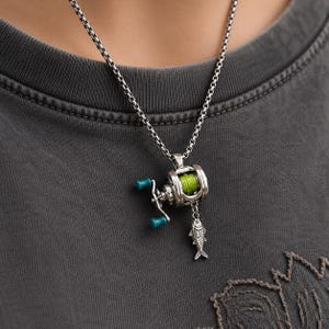 May include: Silver-toned fishing reel pendant necklace with teal accents and a small fish charm. The reel features a green line and is suspended from a chain. The necklace is worn against a gray t-shirt.