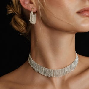 Może przedstawiać: Srebrny naszyjnik z frędzlami i pasujące kolczyki. Naszyjnik jest w stylu choker, z wieloma pasmami delikatnych łańcuszków zwisających w dół. Kolczyki to małe koła z podobnymi detalami frędzli. Biżuteria jest noszona na ciemnym tle.
