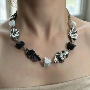 Puede incluir: Un collar plateado con piezas metálicas texturizadas de forma irregular, intercaladas con elementos negros y blancos similares a piedras. El collar tiene un cierre de cadena.