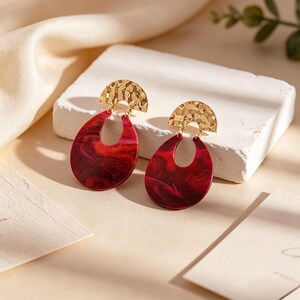 May include: A pair of gold-coloured, textured half-circle earring tops, connected to large, red marbled teardrop-shaped pendants. The earrings are displayed on a white block against a soft, neutral background.