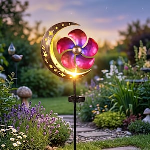 Puede incluir: Una estaca de jardín con energía solar con un diseño de luna creciente y estrellas, y un molinillo de viento con forma de flor. El molinillo de viento tiene pétalos rojos y morados. La estaca está en un jardín con flores y vegetación.