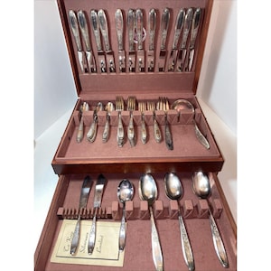 May include: A wooden flatware chest with a hinged lid and a pull-out drawer, lined with mauve fabric. The set includes silver-toned knives, forks, and spoons with ornate handles. A small rectangular card with the text To Keep Silverware Loveliest is included.