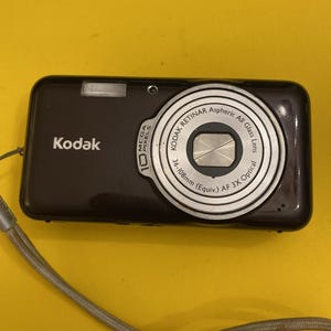 May include: A dark brown Kodak digital camera with a silver lens and flash. The camera has the text "Kodak" and "10 Mega Pixels" printed on it. The lens has the text "KODAK RETINAR Aspheric All Glass Lens" and "AF 3X Optical". A gray wrist strap is attached.