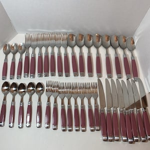 May include: A large flatware set featuring stainless steel utensils with solid mauve-colored handles. The collection includes forks, spoons, and knives arranged in rows, each piece finished with a metallic silver band near the handle base.
