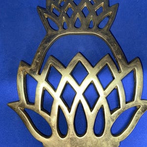 May include: A decorative brass trivet with an openwork design. The trivet features a floral motif with diamond and teardrop shapes. The brass has a slightly aged appearance, and the background is a solid blue.