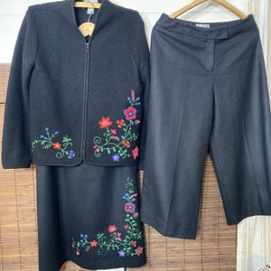 May include: A black three-piece suit featuring a zip-up jacket, a skirt, and wide-leg pants. The jacket and skirt are adorned with colorful floral embroidery in red, blue, and green. The suit is displayed on wooden hangers.