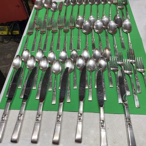 May include: A large collection of silver-toned stainless steel flatware arranged in rows, including various dinner knives with decorative handles, forks, and spoons. The set features multiple pieces of cutlery with polished metal surfaces.