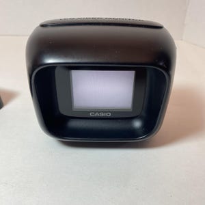 May include: A black Casio LCD video monitor with a rounded, compact housing. The device features a small, rectangular screen centered within a recessed frame, with the text "LCD VIDEO MONITOR" embossed on the top and "CASIO" printed below the screen.