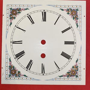 May include: A square, white clock face with black Roman numerals and hands. The face features decorative floral corner accents in shades of brown, pink, and green, with blue and pink border details. Two red dots are centered near the top.