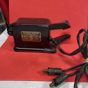 May include: A vintage black sewing motor controller with a metal foot pedal. The controller has a label that reads "Sewing Motor Controller" with technical specifications. The electrical cord and plugs are visible. The item is set against a red background.