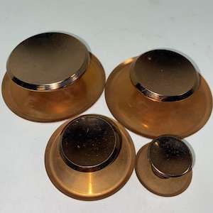 May include: A set of four circular knobs in varying sizes, each featuring a polished metallic copper-toned top surface and a wider, translucent amber-colored base. The knobs are arranged in a cluster, displaying a smooth, reflective finish.