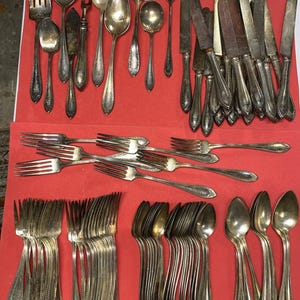 May include: An assortment of antique silverware, including forks, spoons, knives, and a ladle, arranged on a red surface. The silverware is silver-toned with ornate handle designs, showcasing a vintage aesthetic. A collection of dining utensils.