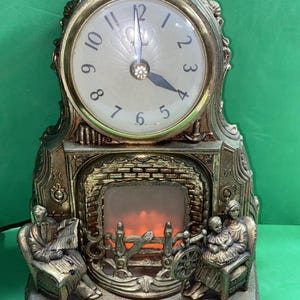 May include: A gold-toned mantel clock featuring a round clock face with black numerals and hands. Below the clock is a decorative fireplace scene with an orange light effect, flanked by two figures sitting in chairs, one reading a newspaper.