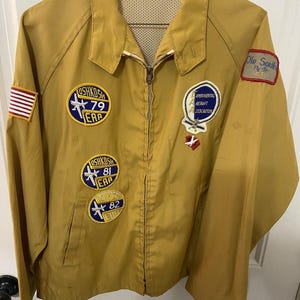 May include: Mustard-colored zip-up jacket with multiple patches. Patches include an American flag, "Oshkosh 79 EAA", "Experimental Aircraft Association", "Ole South Fly-In", "Oshkosh 81 EAA", and "Oshkosh 82 EAA".