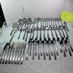 May include: A large collection of stainless steel flatware featuring a decorative pattern on the handles. The set includes various knives, forks, spoons, and serving utensils, accompanied by a yellow note listing the item count.