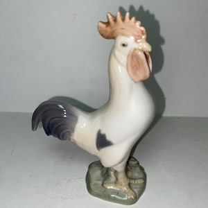 May include: A porcelain rooster figurine featuring a white body with a dark gray tail and a patch on its side. The rooster has a light brown comb and wattle, standing on a textured, light green base. The figure is rendered in a smooth, glossy finish.