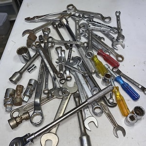 May include: A collection of assorted metal hand tools including various open-end and box-end wrenches, ratchets, socket adapters, and screwdrivers with translucent red, yellow, and blue handles scattered across a flat white surface.