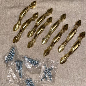 May include: A set of seven gold-toned metal cabinet pulls with a curved, textured design featuring pointed ends and decorative rope-like detailing. The set includes several small plastic bags containing silver-colored metal mounting screws.
