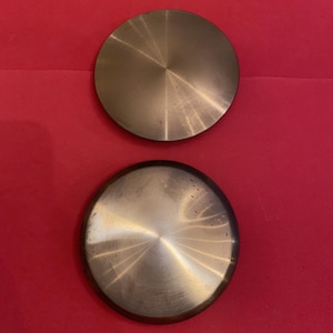May include: Two circular, metallic objects with a brushed finish, arranged against a red background. The objects have a concentric design radiating from the center. The top object is clean, while the bottom one shows slight wear.