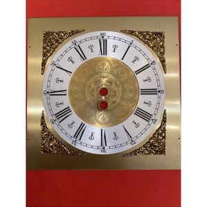 May include: A square brass clock face featuring a circular white dial with black Roman numerals and minute markers. The center is a gold-toned disc with an embossed floral pattern and two red circular openings. Four ornate gold corner spandrels.