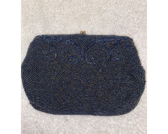 May include: A dark navy blue clutch handbag completely covered in small, iridescent beads. The exterior features a decorative swirling pattern formed by the beadwork across the top section and a small gold-toned metal clasp at the center top.