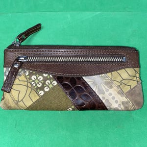 May include: A rectangular wallet featuring a patchwork design of olive green, brown, and beige fabric panels with floral and geometric patterns. It includes a dark brown leather top section, a zippered front pocket, and a main zipper closure.