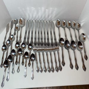 May include: A large collection of silver-toned stainless steel flatware including various spoons, forks, and knives. Each piece features a decorative embossed pattern on the handle, with polished bowls and tines arranged in a grid-like formation.