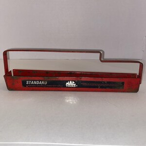 May include: A red metal Mac Tools socket rail organizer featuring a long handle and a black label with the text STANDARD and MAC TOOLS. The tray shows signs of wear and rust along the edges, with safety warnings printed on the front label.
