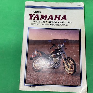 May include: A Clymer Yamaha XV535-1100 Virago 1981-1997 service, repair, and maintenance manual. The white cover features a photograph of a motorcycle on a dry, cracked landscape, with the text YAMAHA and model information printed in black and purple.