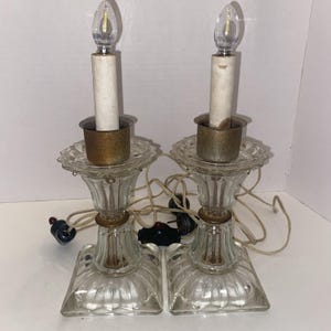 May include: A pair of electric candle lamps featuring clear, ribbed glass bases with square, faceted feet. Each lamp has a brass-toned metal bobeche, a white faux-candle sleeve, and a clear flame-shaped LED bulb with a visible white power cord.