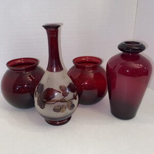 May include: A collection of four glass vessels including two deep red spherical bowls, one tall slender vase with a frosted base featuring etched acorn and leaf designs, and one dark red bottle-shaped vase with a rounded neck and rim.