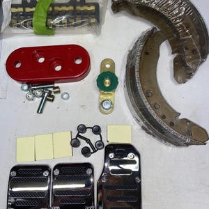 May include: A collection of automotive parts including two curved brake shoes wrapped in plastic, a red oval mounting plate, a green and gold metal component, three chrome-finished pedal covers with black textured inserts, small hardware, and adhesive pads.