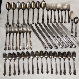 May include: A large set of silver-toned flatware featuring ornate floral detailing on the handles. The collection includes numerous spoons, forks, and knives arranged in rows, showcasing a consistent decorative pattern across all individual pieces.