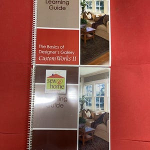 May include: Two spiral-bound sewing guides with red and brown covers. The top guide reads "Learning Guide" and "The Basics of Designer's Gallery Custom Works II." The bottom guide reads "Learning Guide" and "Basic Computer Skills for Embroiderers."