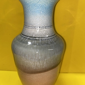 May include: A ceramic vase with a gradient design. The top of the vase is light blue, transitioning to white, then gray, and finally a tan color at the base. The vase has a flared top and a rounded body.