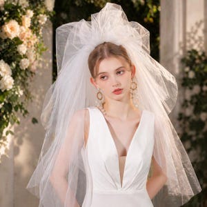 Puede incluir: Una novia con un vestido de novia blanco con escote en V profundo y un velo transparente. El velo está sujeto a un tocado y cae sobre los hombros. La novia lleva pendientes dorados con perlas.