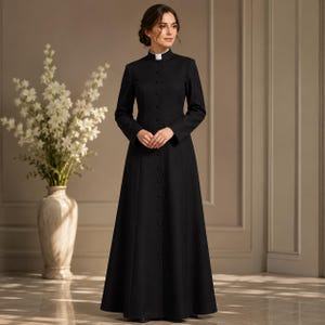 Puede incluir: Un vestido sotana negro hasta el suelo con mangas largas, cuello alto tipo mandarín con una inserción blanca y una tapeta de botones frontal completa con botones negros a juego. La prenda tiene un corpiño ajustado y una falda acampanada.