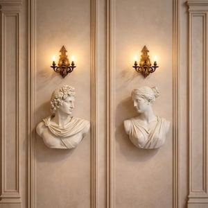 Könnte beinhalten: Zwei weiße Marmorbüsten klassischer Figuren sind an einer beigefarbenen Wand angebracht, flankiert von verzierten goldenen Wandleuchtern mit brennenden Kerzen. Die Büsten zeigen einen Mann und eine Frau, jeweils mit detaillierten Gesichtszügen und drapierten Kleidungsstücken. Die Wand hat vertikale Paneele.