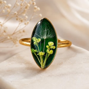 May include: A gold-toned ring featuring a marquise-shaped resin setting. The resin is deep green and contains embedded yellow flowers and a single white dandelion seed head. The band is thin and polished, creating a sleek and simple appearance.