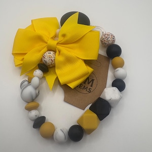 May include: A yellow bow with a variety of beads in black, white, and yellow. The beads are in various shapes, including round and cube. A tag with the letters "EMS" is attached.