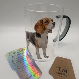 May include: A clear glass mug featuring a beagle dog illustration. The mug has a dark grey handle and a care instruction tag. A brown tag with the text "TM GEMS" is also present.