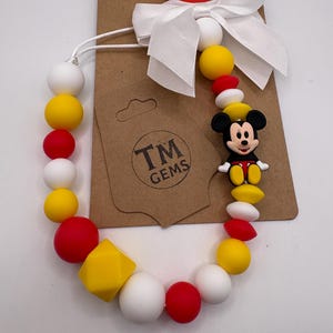 May include: A dummy clip featuring a red plastic clasp, a white satin ribbon bow, and a string of silicone beads in red, yellow, and white. The beads include round and faceted shapes, with a Mickey Mouse character charm and a TM GEMS logo tag.