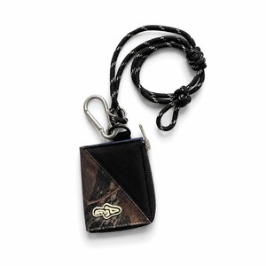 Könnte beinhalten: Schwarzes und Camouflage-Muster-Portemonnaie mit silbernem Reißverschluss und einem schwarz-weißen Seilband. Das Portemonnaie hat einen silbernen Karabinerhaken und ein kleines silbernes Emblem auf der Vorderseite.